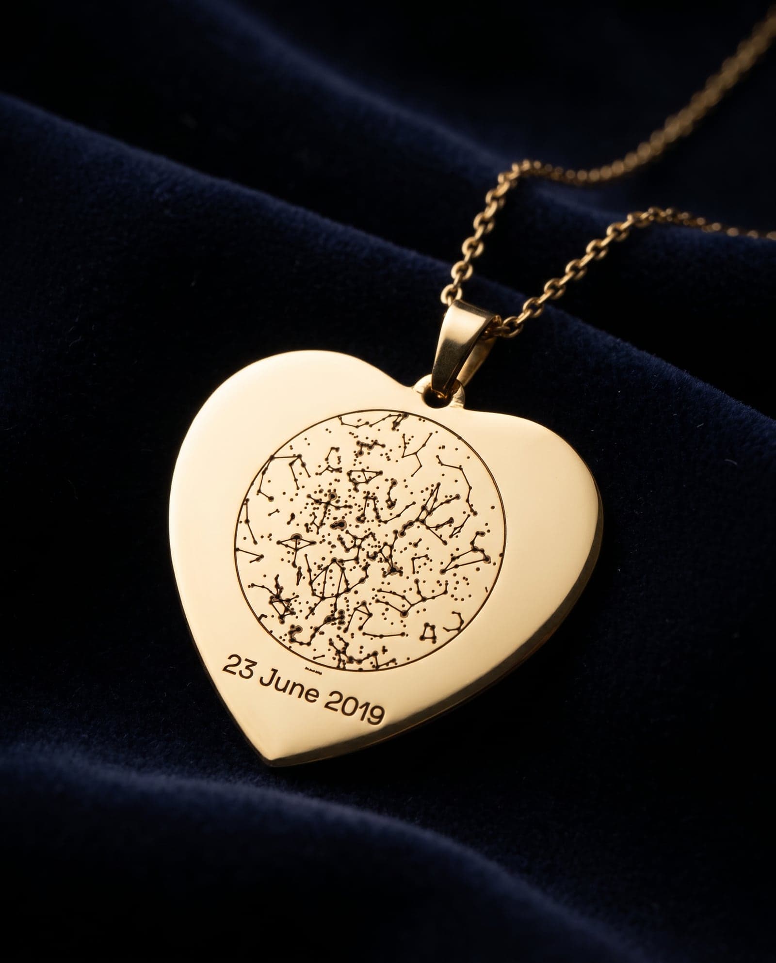 A gold heart pendant engraved with a star chart and a date, on navy velvet