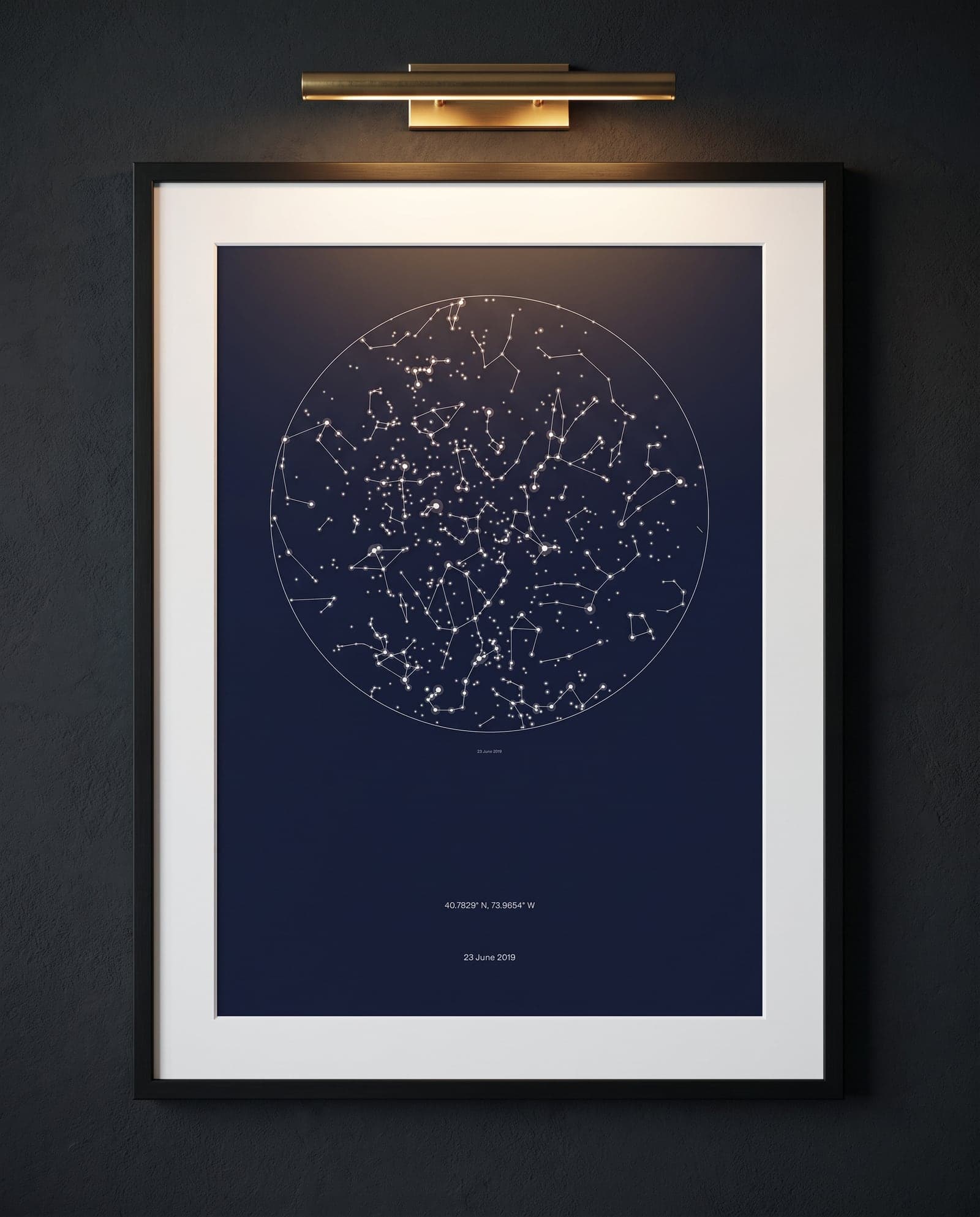 A navy star chart framed behind glass, lit by a picture light on a dark wall